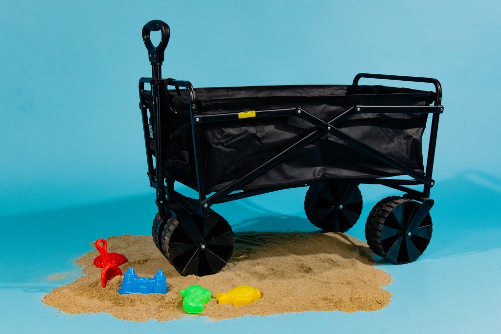 Black collapsible wagon with its front wheels on a pile of sand, with children's multicolor plastic beach toys.