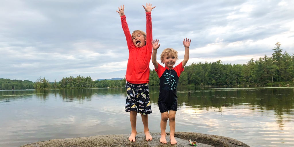 Two children wearing swim gear, standing side by side in front of a body of water.